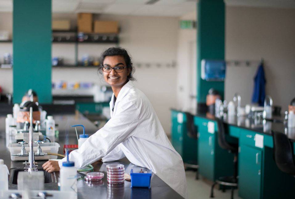 Student in white lab coat smiles into the camera as they work in a lab